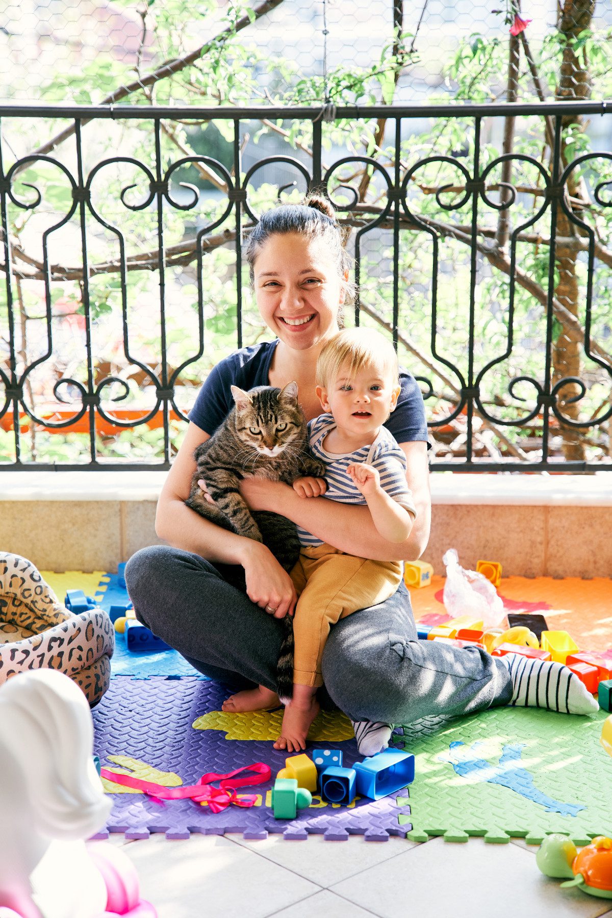 Mom sits on a rug on the balcony and holds a small child and a cat on her lap
